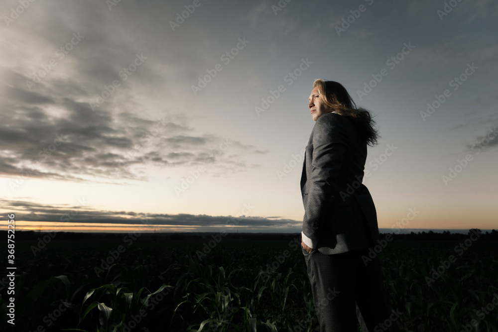 Fototapeta premium young businessman stands in a field in a gray suit at sunset. Blond with long hair in a cornfield