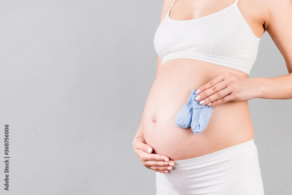Close up portrait of pregnant woman in white underwear holding blue socks for a baby boy at gray background. Future child expectation concept. Copy space Close up portrait of pregnant woman in white underwear holding blue socks for a baby boy at gray background. Future child expectation concept. Copy space