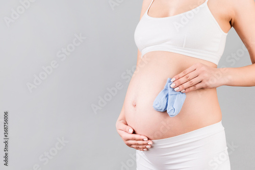 Close up portrait of pregnant woman in white underwear holding blue socks for a baby boy at gray background. Future child expectation concept. Copy space