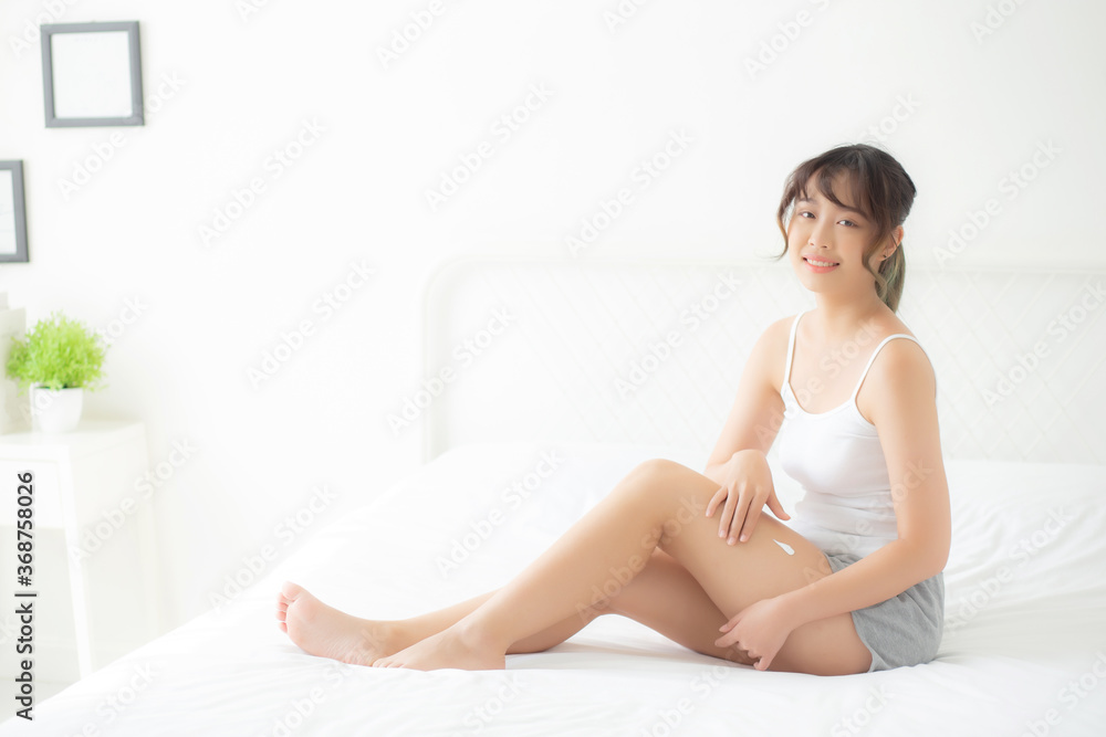 Beautiful young asian woman sitting on a bed stroking legs with soft smooth skin in the bedroom, girl applying touch body cream and lotion with treatment care, healthy and wellness concept.