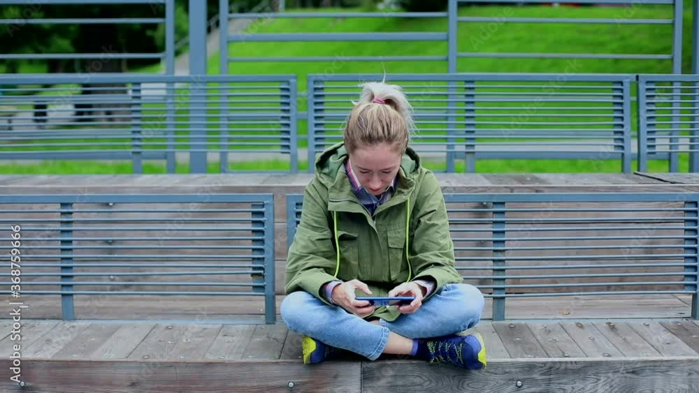 Stylish girl gaming online on smartphone mobile resting on sports area outside. Female gamer playing with friends. Social networking. Internet surfing. Tapping touchscreen phone. Sunny day light. Stylish girl gaming online on smartphone mobile resting on sports area outside. Female gamer playing with friends. Social networking. Internet surfing. Tapping touchscreen phone. Sunny day light.