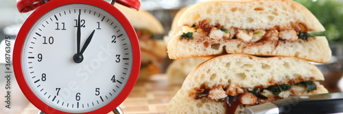 Close-up of red clock standing on wooden table and showing one pm on dial. Sandwiches and sharp knife. Lunch time. Break from work. Fast food delivery and nutrition concept