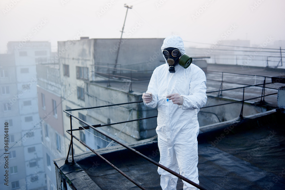 Scientist wearing white protective coverall, gloves and gas mask, holding in his hands tweezers and glass flask with blue reagent, block of flats on background