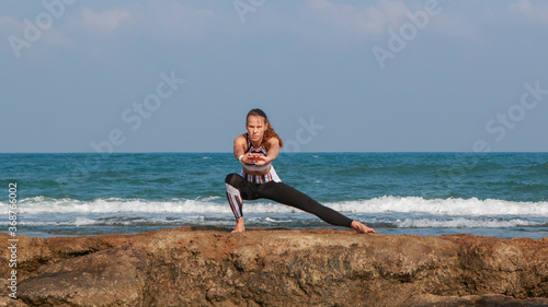 Thin athletic woman in a stretching pose, on beach rocks with the sea behind her.
