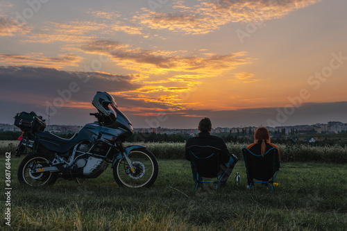 Couple of motobikers tourists sit on folding chairs and look to distance, ejoy the sunset view. Escape from the city. Outdoors nature. Motorcycle road trip. Freedom. Copy space photo