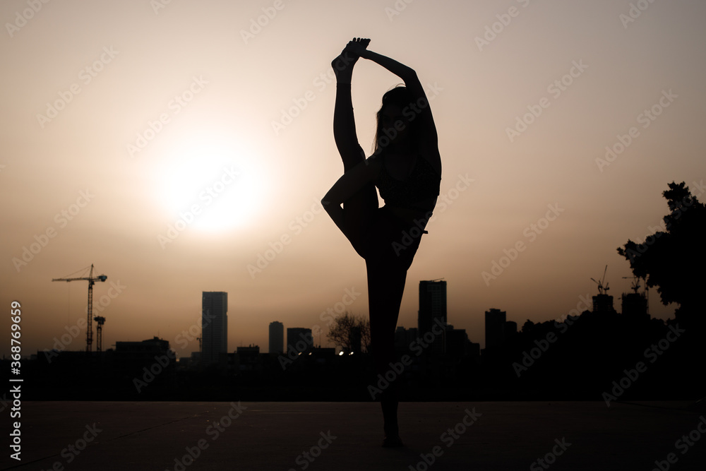 Flexible young woman stretching in an empty urban park. Silhouette shot with Tel Aviv buildings in far background.