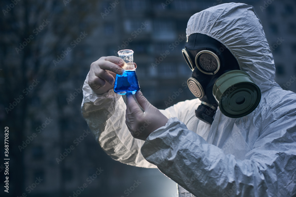 Fototapeta premium Close-up view, scientist wearing white coverall and a gas mask, holding glass flask with samples, worker observing reaction on the light, high building on background