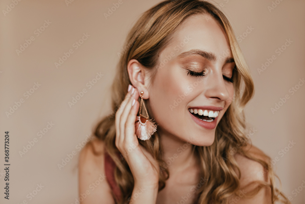 Cheerful young lady touching her earring with smile. Indoor shot of ecstatic laughing girl isolated on light background. Cheerful young lady touching her earring with smile. Indoor shot of ecstatic laughing girl isolated on light background.