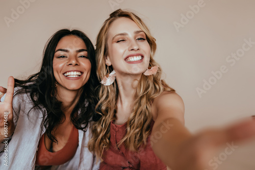 Excited blonde woman with glamorous accessories making selfie. Indoor photo of ecstatic girls funny posing in studio.