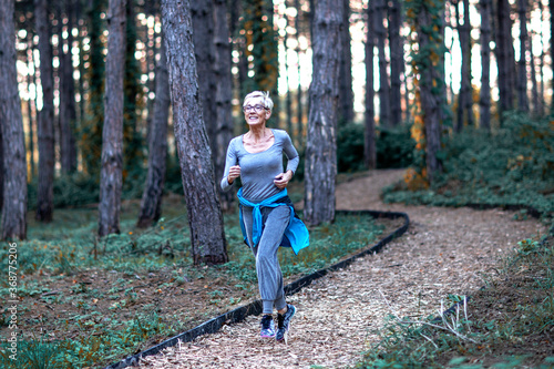 Aged woman jogging in park between woods at the end of day and smile