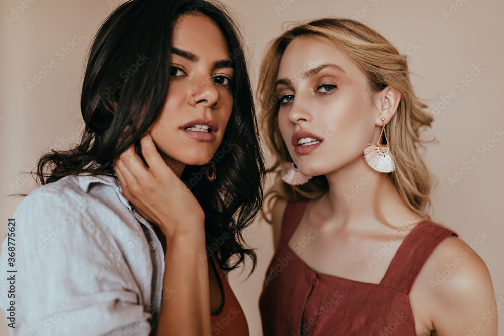 Enchanting brunette girl posing with blonde sister. Serious fashionable women standing on beige background. Enchanting brunette girl posing with blonde sister. Serious fashionable women standing on beige background.