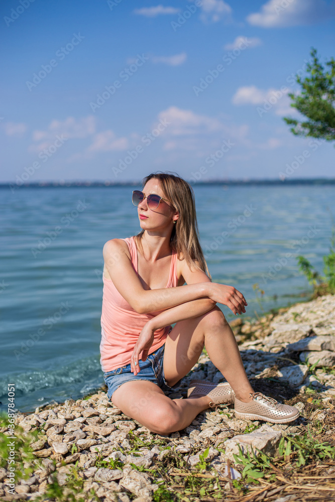 A girl sits on a rocky lake shore on a sunny day