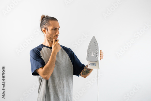 Hombre con moño y camiseta gris de mangas azul oscuro duda de cómo se utiliza una plancha, mientras la sostiene con la mano y la mira confuso