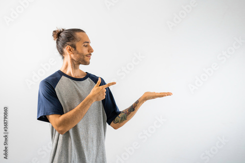Hombre con moño, caucásico, con camiseta gris de mangas y cuello azul oscuro, señala hacia algo que flota sobre su mano. Inserte su logotipo, objeto o texto