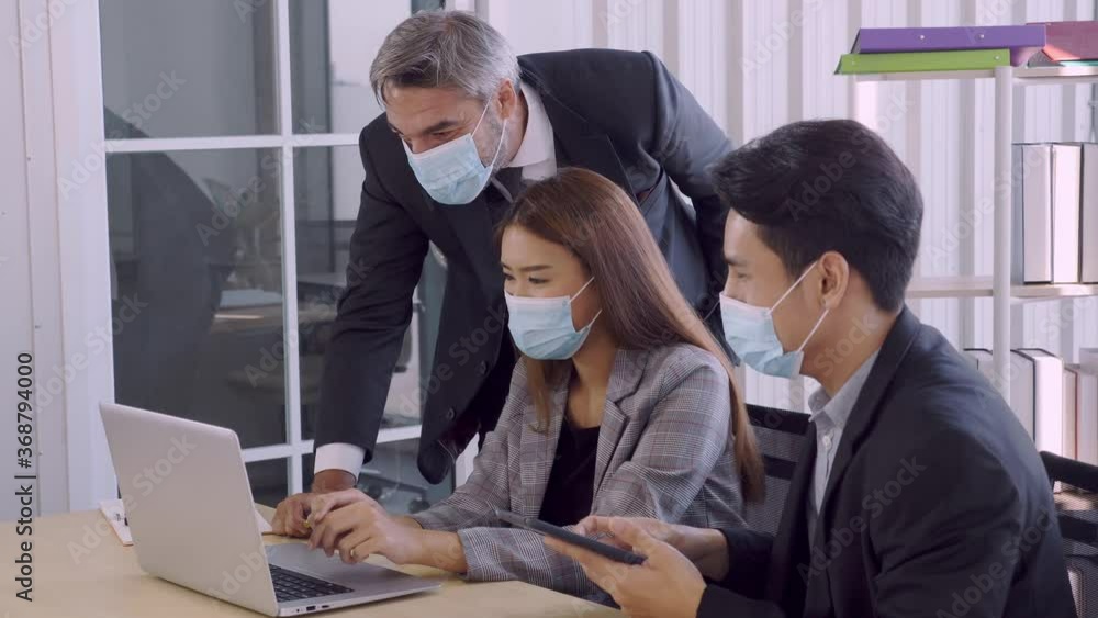 Normal view, medium shot of sernior executive caucasian boss pointing at laptop screen and nod to 2 young asian employee. Everyone wears facial mask during work at office. Normal view, medium shot of sernior executive caucasian boss pointing at laptop screen and nod to 2 young asian employee. Everyone wears facial mask during work at office.
