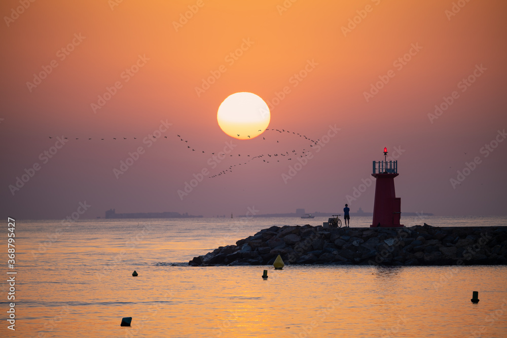 Sunrise at Guardamar jetty in Spain. A cyclist looks at the sea from the lighthouse with the island of Tabarca in the background. A flock of birds in the sky.
