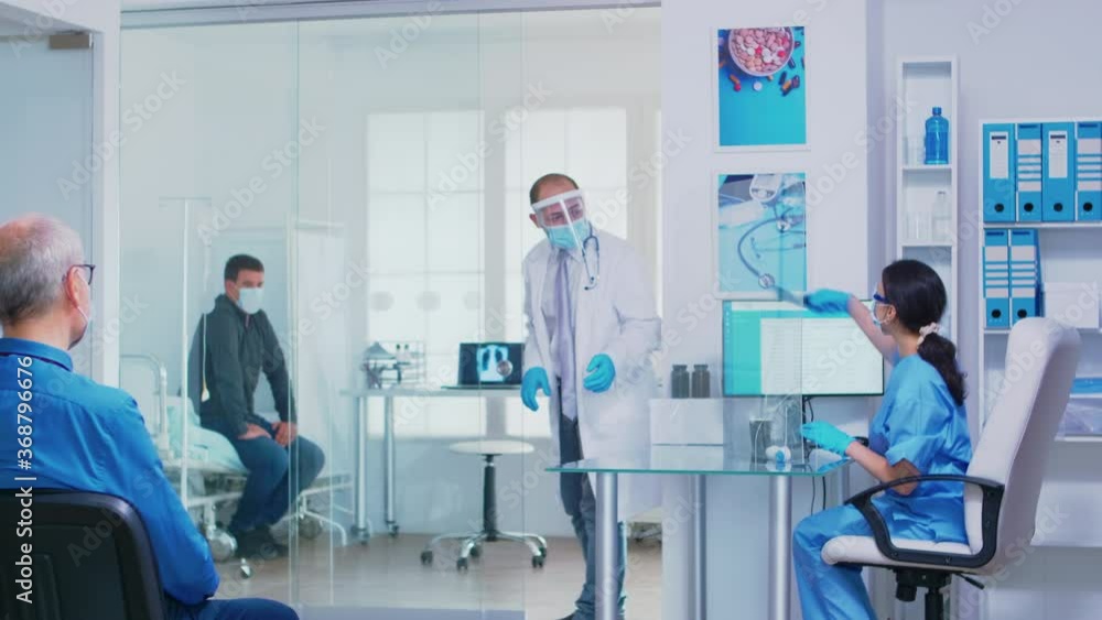 Nurse giving doctor patient radiography wearing face mask against coronavirus. Assistant with visor pusing disabled senior woman in wheelchair through waiting area. Nurse giving doctor patient radiography wearing face mask against coronavirus. Assistant with visor pusing disabled senior woman in wheelchair through waiting area.
