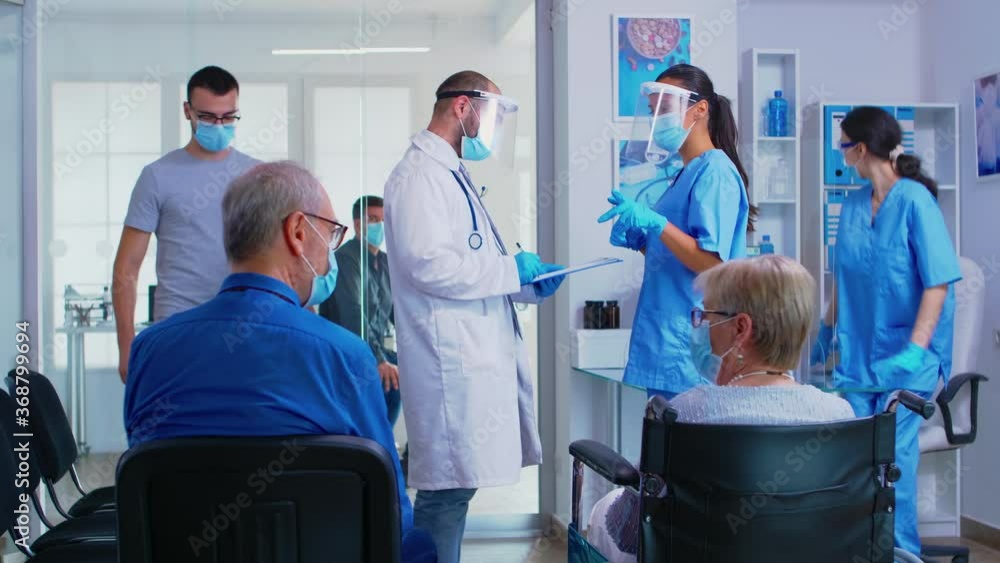 Medical team with face mask against coronavirus discussing in hospital waiting room about patient diagnosis. Senior man talking with disabled woman in wheelchair. Patient waiting for examination. Medical team with face mask against coronavirus discussing in hospital waiting room about patient diagnosis. Senior man talking with disabled woman in wheelchair. Patient waiting for examination.