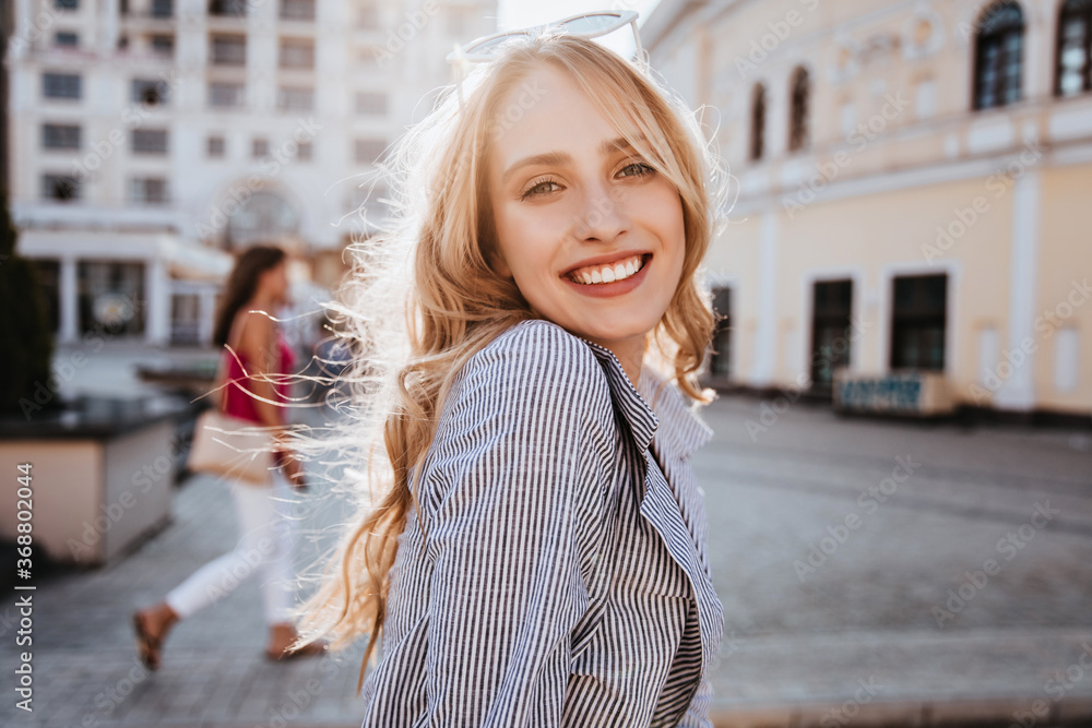 Emotional blonde woman posing with happy smile on the street. Debonair caucasian girl enjoying photoshoot on city background. - obrazy, fototapety, plakaty 