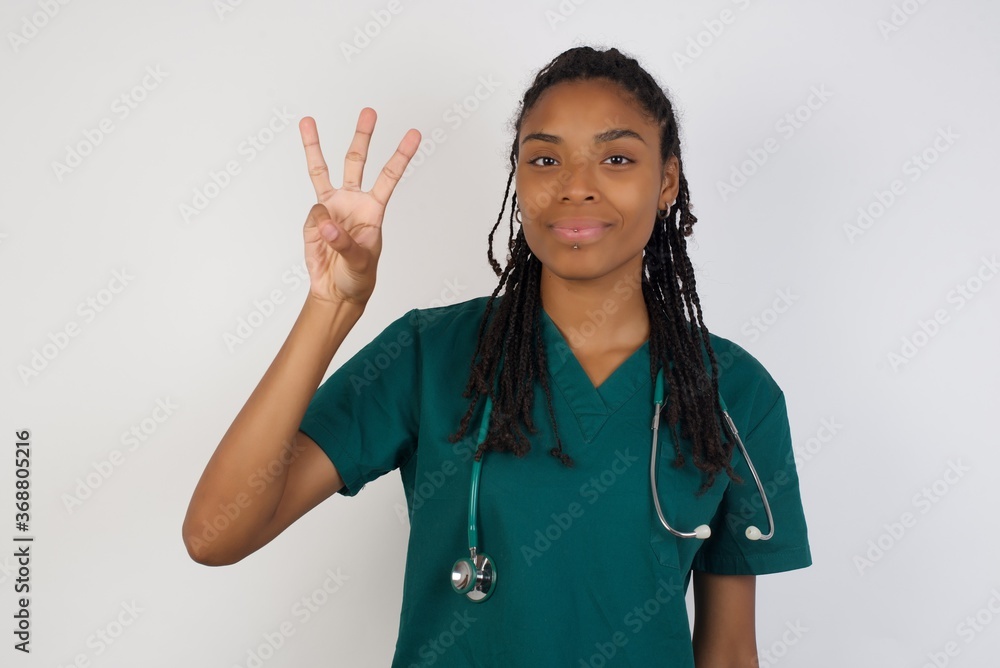 Young doctor woman wearing medical uniform standing against gray wall showing and pointing up with fingers number three while smiling confident and happy. Young doctor woman wearing medical uniform standing against gray wall showing and pointing up with fingers number three while smiling confident and happy.
