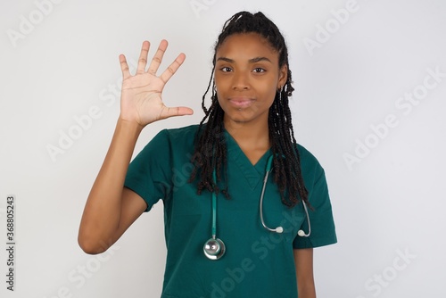 Young doctor woman standing against gray wall showing and pointing up with fingers number five while smiling confident and happy.