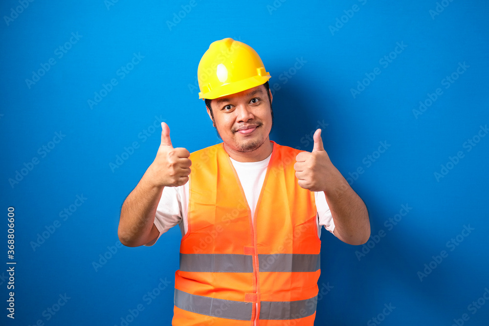 Fat asian construction worker wearing orange safety vest and helmet showing thumb up sign