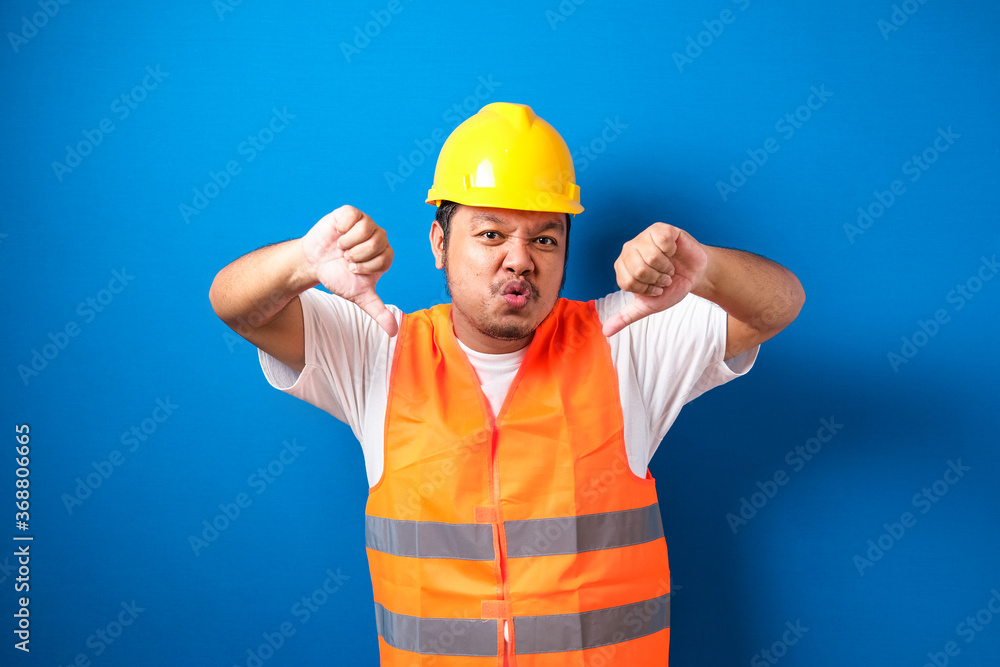 Young fat asian construction worker man wearing orange safety vest and helmet looking unhappy and angry