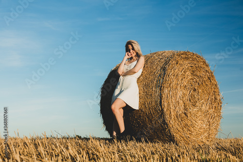 blonde girl is staying by a rolled haystack in field in sunset time