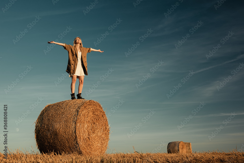 blonde girl is sitting on a rolled haystack in field in sunset time blonde girl is sitting on a rolled haystack in field in sunset time