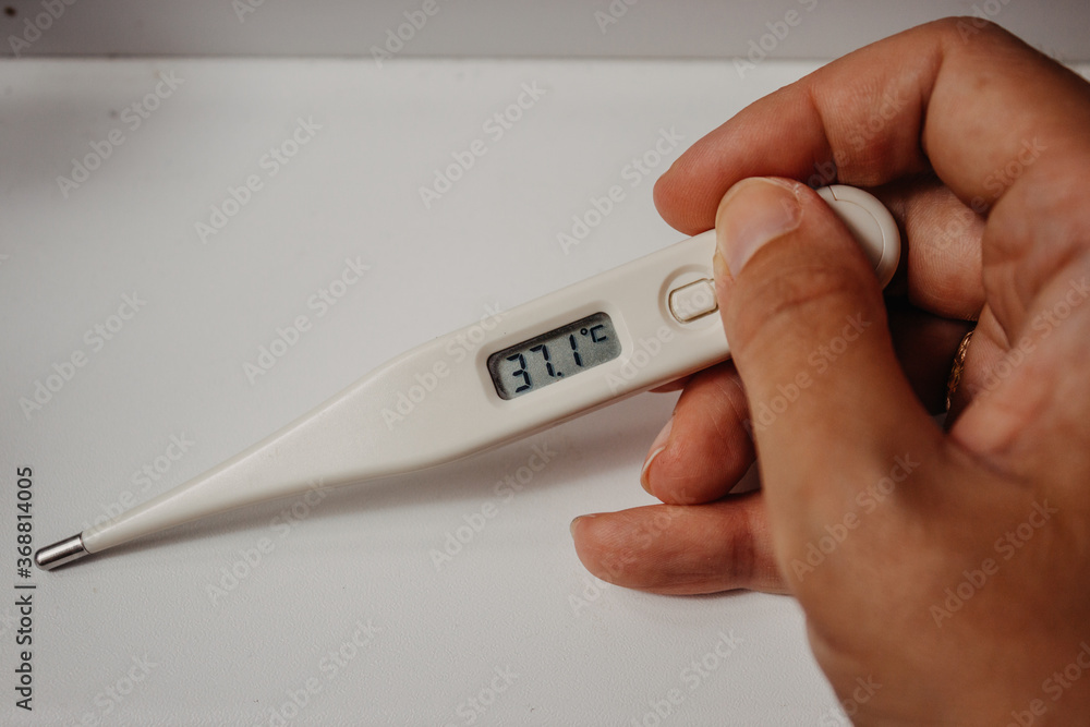 Obraz premium Disease and fever. First symptoms of the coronavirus. Electronic thermometer with a display value of 37.1 degrees in the hand of a woman on a light white background close-up with place for text.