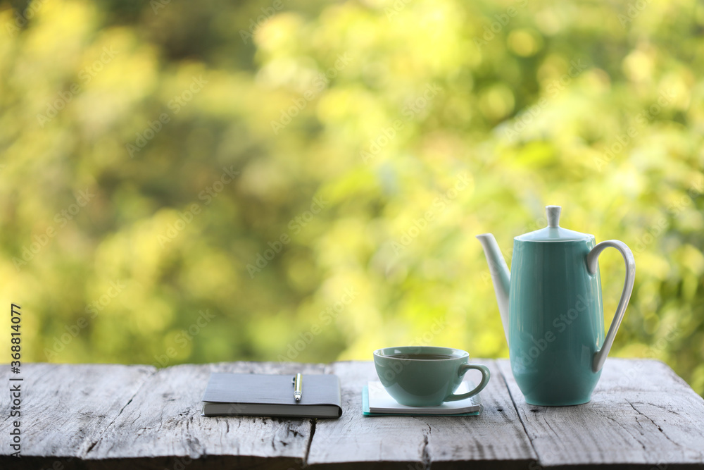 Teal teapot and cup with black book on rustic wooden table with green leaves background Teal teapot and cup with black book on rustic wooden table with green leaves background