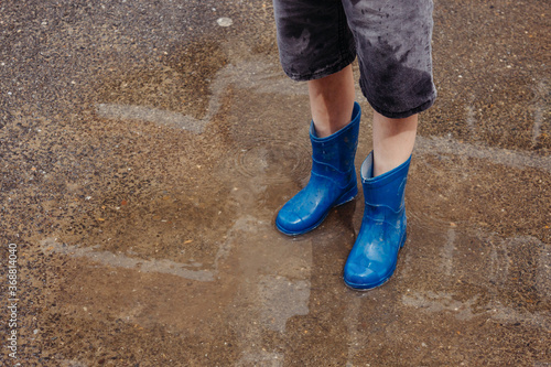 Children in the summer after the rain. Close-up of a boy's feet in blue rubber boots standing in a puddle on a walk. Children walk in the open air after the rain. Waterproof shoes for boys.