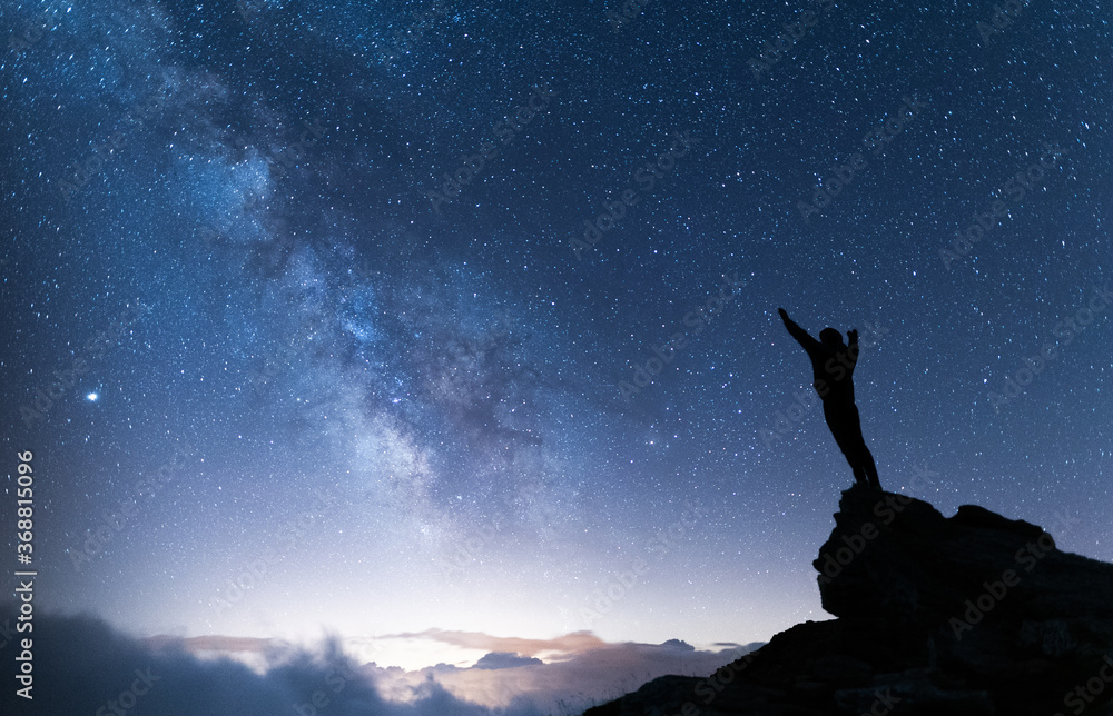Fototapeta premium Person with arms in the air contemplating the vastness of the universe. Silhouette of a man under the milky way and starry magical sky. Real outdoor adventure and wild life concept.