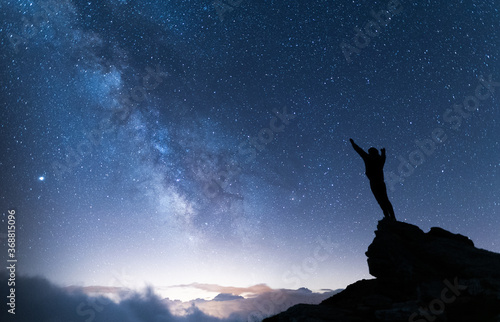 Person with arms in the air contemplating the vastness of the universe. Silhouette of a man under the milky way and starry magical sky. Real outdoor adventure and wild life concept.