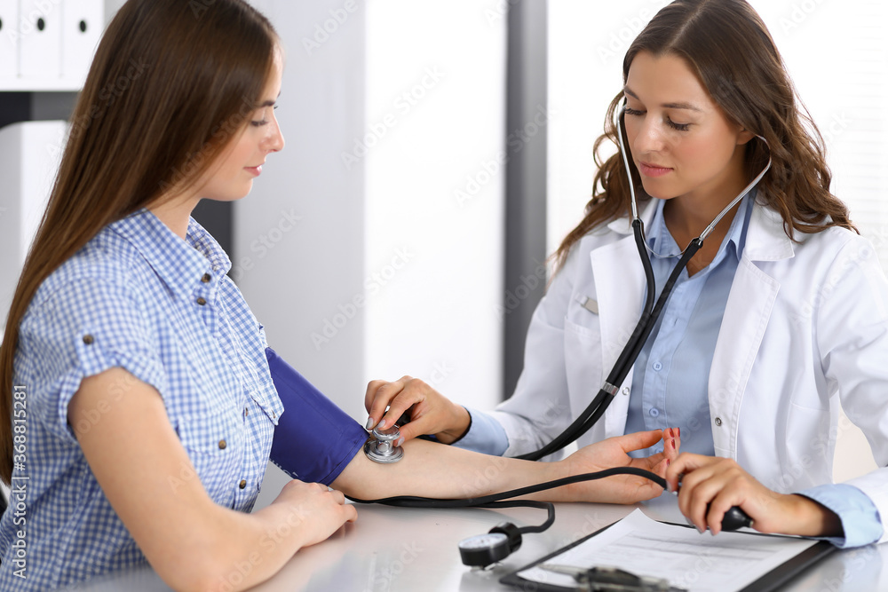 Doctor checking blood pressure of female patient while sitting at the desk in hospital office. Cardiology in medicine and health care concept Doctor checking blood pressure of female patient while sitting at the desk in hospital office. Cardiology in medicine and health care concept
