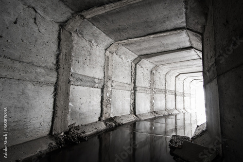 A square concrete drainage tunnel with a light at the end that shines from the turn of the tunnel.