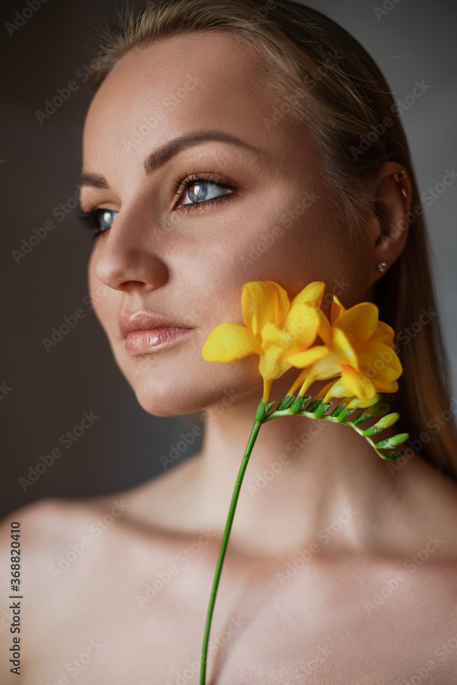 Portrait of a beautiful blonde with blue-green eyes and a yellow freesia flower in her hair. A young woman holds a flower in front of her face. The concept of cosmetology, clean skin, natural beauty