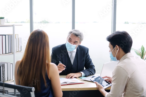 Smart Caucasian senior manager and businessman in black suit talking with asian teamwork staff is wearing masks prevent covid 19 virus at office. New nomal and social distace of business concept. photo