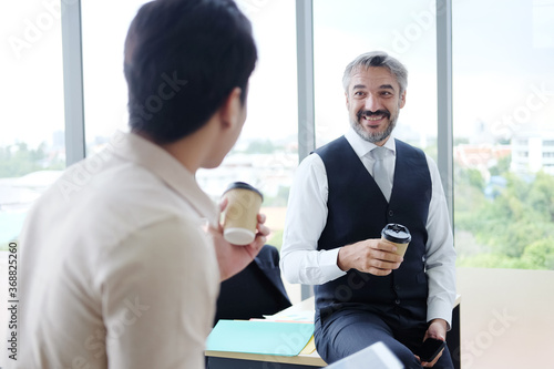 Smiling young Asian and Senior Caucasian businesspeople teamwork relaxing and drinking a mug of hot coffee together at modern office. Business and lifestyle concept.