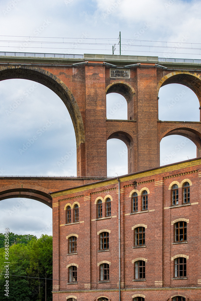 Fototapeta premium Mylau, Saxony, Germany. The building of the historic Ketzel flour mill is dwarfed by the arches of the Goeltzschtalbridge, the largest brick-built bridge in the world,