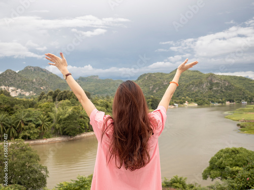 Portrait of happy Asian woman in pink dress standing with river, mountain, and sky in background at Kanchanaburi, Thailand.