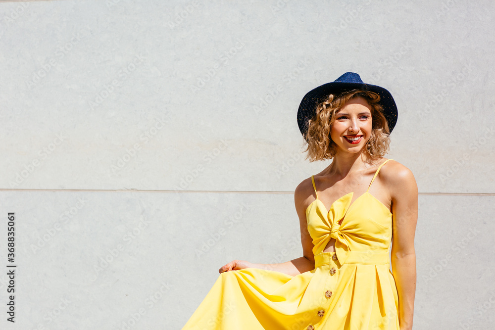 Fototapeta premium Portrait of a smiling young woman with summer hat and trendy yellow dress standing outside