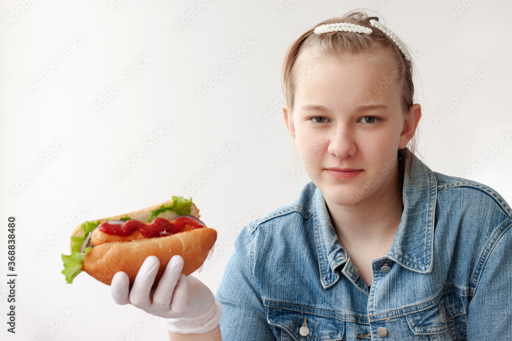 A young girl with blond hair in a blue denim jacket and medical gloves holds a hot dog and looks at the camera