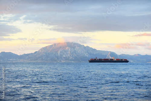 Cargo ship sailing near the Rock of Gibraltar at sunrise, a view from the yacht. British Overseas Territory. Epic cloudscape. Travel destinations, logistics, global communications photo