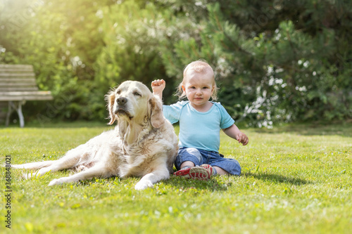 Cute liitle baby boy is waving at the camera posing with Golden Retriever dog in the garden
