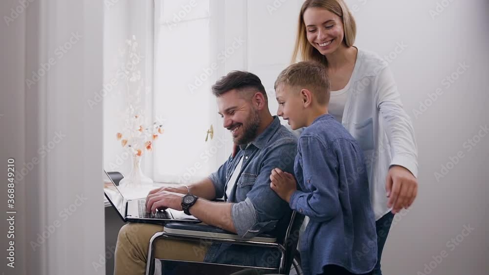 Likable smiling 8-aged boy approaching to his satisfied cheerful young mother and happy bearded dad which sitting in wheelchair and working on computer near window Likable smiling 8-aged boy approaching to his satisfied cheerful young mother and happy bearded dad which sitting in wheelchair and working on computer near window