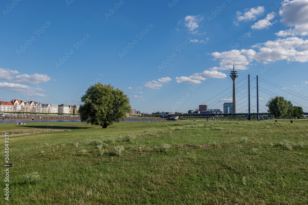 Outdoor sunny scenery of natural promenade and green field on riverside of Rhine River, and background of skyline cityscape of Düsseldorf city and Rhine Tower, Germany in summer season. Outdoor sunny scenery of natural promenade and green field on riverside of Rhine River, and background of skyline cityscape of Düsseldorf city and Rhine Tower, Germany in summer season.
