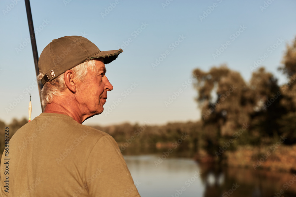 Side view of senior man sitting on bank of river and looking aside, having rest in open air, enjoying beautiful nature near lake, wearing green t shirt and cap.