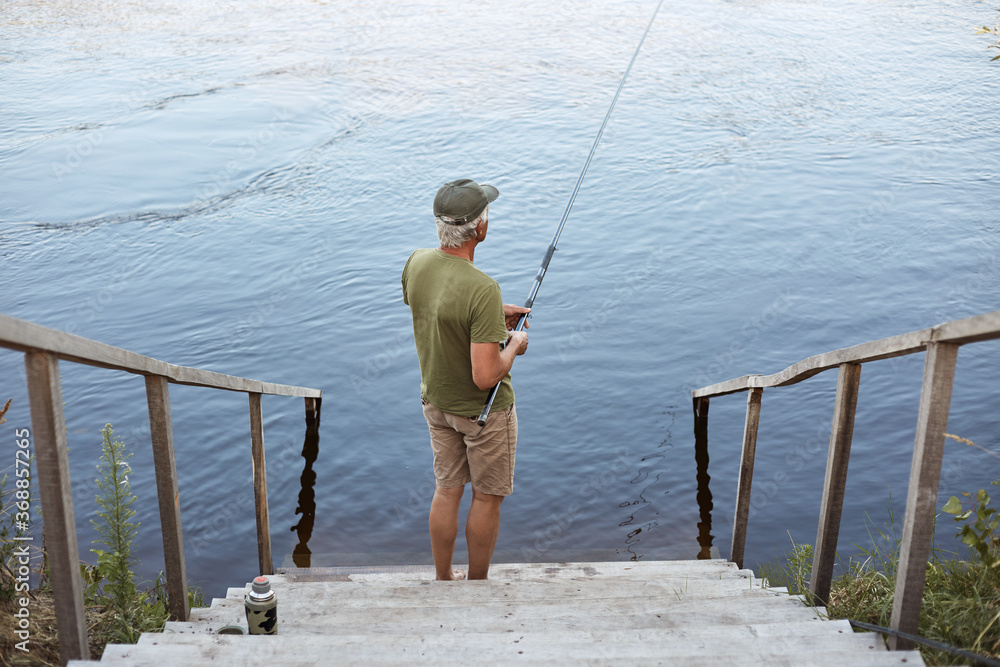 Backwards view of man catching fish while posing on wooden stairs leading to lake, male wearing casual attire, enjoying beautiful nature and fishing. Backwards view of man catching fish while posing on wooden stairs leading to lake, male wearing casual attire, enjoying beautiful nature and fishing.
