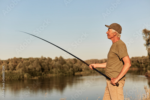Fisherman using rod fly fishing in river, standing at bank of lake with concentrated look, catching fish, wearing casual clothing, spends time outdoors.